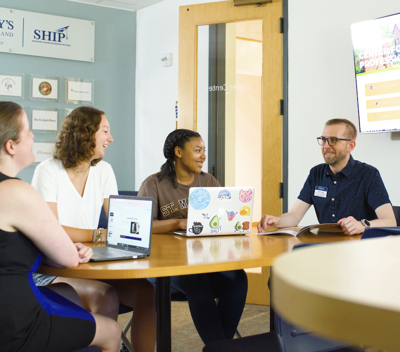 Four people are sitting around a table in an office setting. They are engaged in conversation, with two using laptops. A screen is visible on the wall displaying text. The atmosphere appears collaborative.